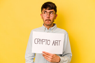 Young hispanic man holding crypto art placard isolated on yellow background confused, feels...