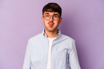 Young caucasian man isolated on purple background blows cheeks, has tired expression. Facial expression concept.