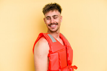 Young caucasian man wearing a life jacket isolated on yellow background looks aside smiling,...