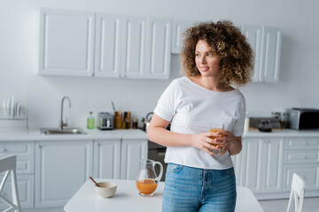 curly woman with glass of fresh orange juice smiling in blurred kitchen.