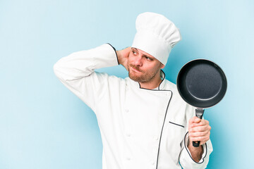 Young caucasian chef man holding flying pan isolated on blue background touching back of head, thinking and making a choice.