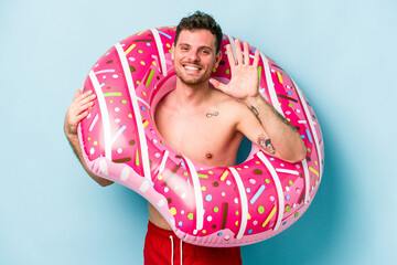 Young caucasian man holding an inflatable donut isolated on blue background smiling cheerful...