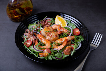 Salad with seafood, arugula, tomatoes, cucumbers, red onion and lemon on a black plate. Black background, close-up.