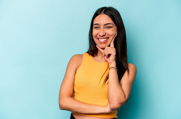 Young hispanic woman isolated on blue background smiling happy and confident, touching chin with...