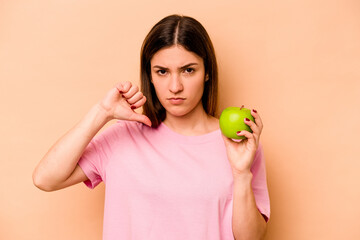 Young hispanic woman holding an apple isolated on beige background showing a dislike gesture, thumbs down. Disagreement concept.