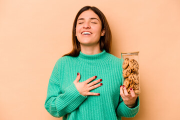 Young hispanic woman holding cookies jar isolated on beige background laughs out loudly keeping...