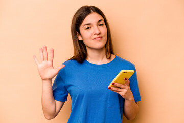 Young caucasian woman holding mobile phone isolated on beige background smiling cheerful showing number five with fingers.