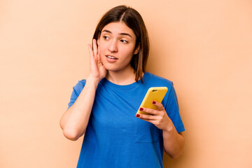 Young caucasian woman holding mobile phone isolated on beige background trying to listening a gossip.