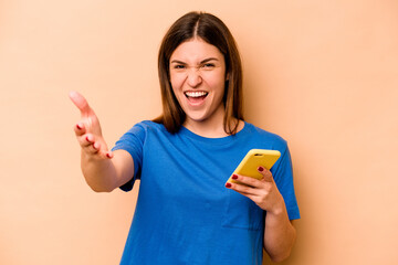 Young caucasian woman holding mobile phone isolated on beige background receiving a pleasant surprise, excited and raising hands.