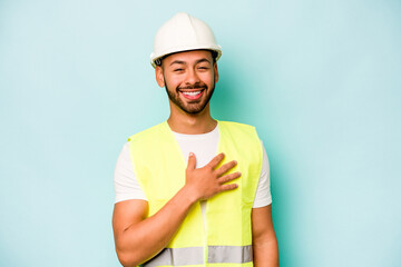 Young laborer hispanic man isolated on blue background laughs out loudly keeping hand on chest.