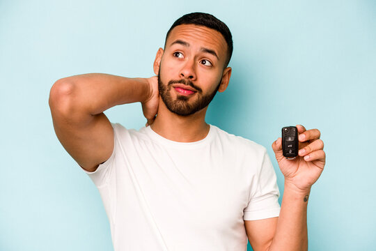 Young hispanic man holding car keys isolated on blue background touching back of head, thinking and making a choice.