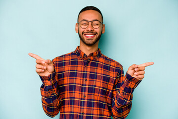 Fototapeta premium Young hispanic man isolated on blue background pointing to different copy spaces, choosing one of them, showing with finger.