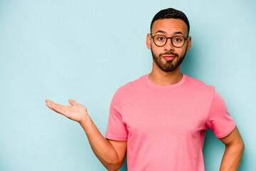 Young hispanic man isolated on blue background showing a copy space on a palm and holding another...