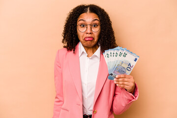 Young African American business woman holding banknotes isolated on beige background shrugs shoulders and open eyes confused.