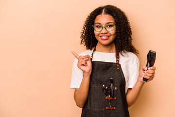 Young African American hairdresser woman isolated on beige background smiling and pointing aside,...