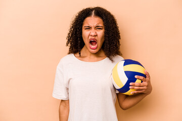 Young African American woman playing volleyball isolated on beige background screaming very angry...