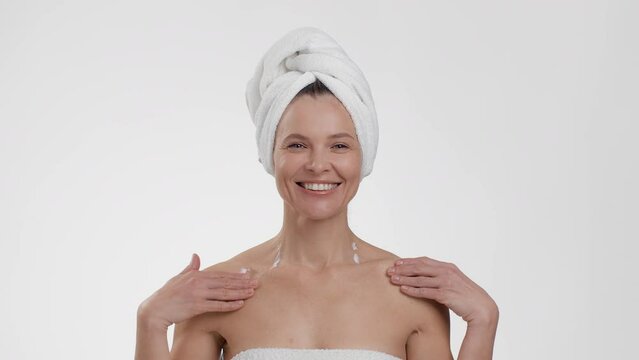 Beautiful happy middle aged woman pampering her body after shower with pampering lotion, posing with towel on head