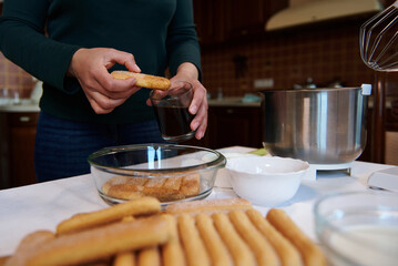 Cropped image of dipping biscuit cookies into a glass with strong sweet espresso coffee while making traditional Italian dessert Tiramisu in vintage home kitchen