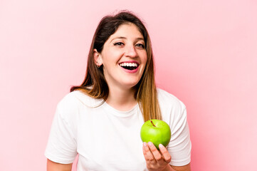 Young caucasian woman holding an apple isolated on pink background laughing and having fun.