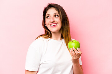 Young caucasian woman holding an apple isolated on pink background looks aside smiling, cheerful and pleasant.