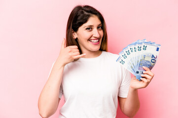 Young caucasian woman holding banknotes isolated on pink background showing a mobile phone call gesture with fingers.