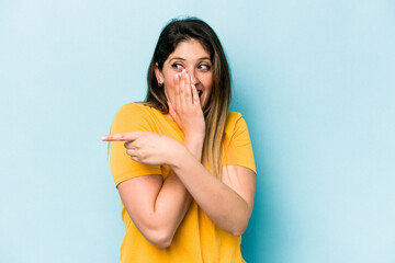 Young caucasian woman isolated on blue background saying a gossip, pointing to side reporting something.