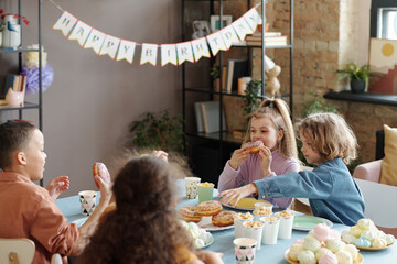 Group of children sitting at table and enjoying donuts and other sweet food during birthday party...