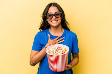 Young hispanic woman holding popcorn isolated on yellow background laughs out loudly keeping hand on chest.