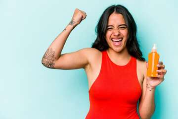 Young hispanic woman holding sun cream isolated on blue background raising fist after a victory, winner concept.