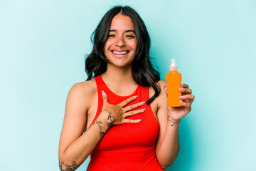 Young hispanic woman holding sun cream isolated on blue background laughs out loudly keeping hand on chest.