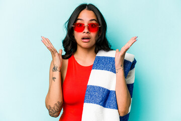 Young hispanic woman holding beach towel isolated on blue background surprised and shocked.