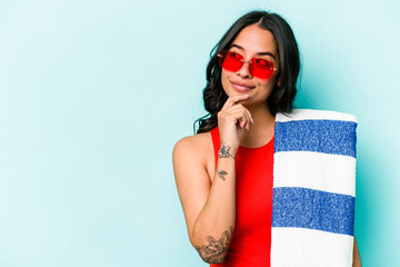 Young hispanic woman holding beach towel isolated on blue background looking sideways with doubtful...