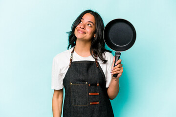 Young hispanic cooker woman holding frying pan isolated on blue background dreaming of achieving...
