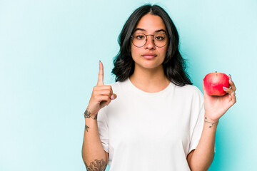 Young hispanic woman holding an apple isolated on blue background showing number one with finger.