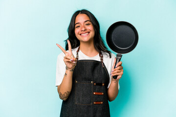 Young hispanic cooker woman holding frying pan isolated on blue background showing number two with...