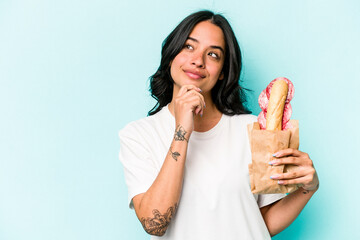 Young hispanic woman eating a sandwich isolated on blue background looking sideways with doubtful...