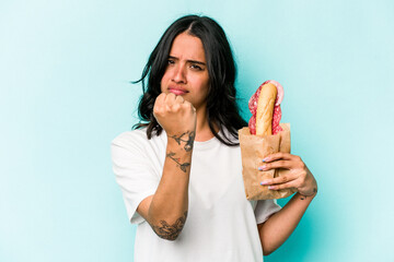 Young hispanic woman eating a sandwich isolated on blue background showing fist to camera, aggressive facial expression.