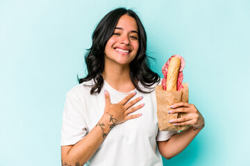 Young hispanic woman eating a sandwich isolated on blue background laughs out loudly keeping hand on chest.
