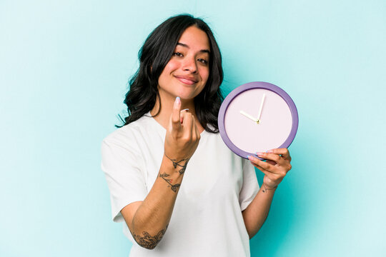 Young hispanic woman holding a clock isolated on blue background pointing with finger at you as if inviting come closer.