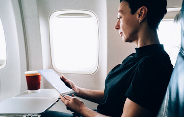 Brunette 40 years old female airplane passenger with tablet in hands sitting in comfortable seat near the window. Attractive woman enjoying flight while using with touch pad online connection on board