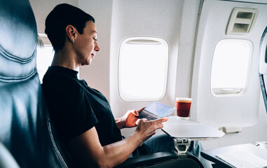 Brunette 40 years old female airplane passenger with tablet in hands sitting in comfortable seat near the window. Attractive woman enjoying flight while using with touch pad online connection on board