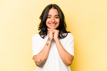 Young hispanic woman isolated on yellow background keeps hands under chin, is looking happily aside.