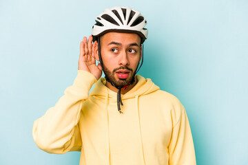 Young hispanic man wearing helmet isolated on blue background trying to listening a gossip.
