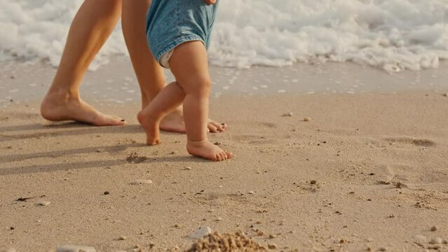 Little baby learns to walk. Close up on feet. Sea sunset. Mother is support her child to do the first steps on beach sand in summer. Toddler is learning to walk, Slow Motion