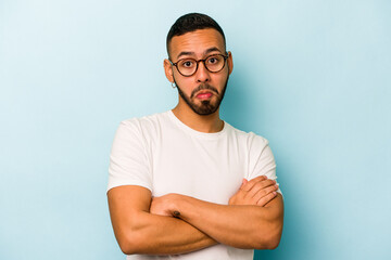 Young hispanic man isolated on blue background shrugs shoulders and open eyes confused.