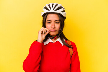 Young student hispanic woman wearing a bike helmet isolated on yellow background with fingers on lips keeping a secret.