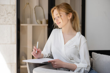 Blonde woman in blouse holding pen and notebook at home.