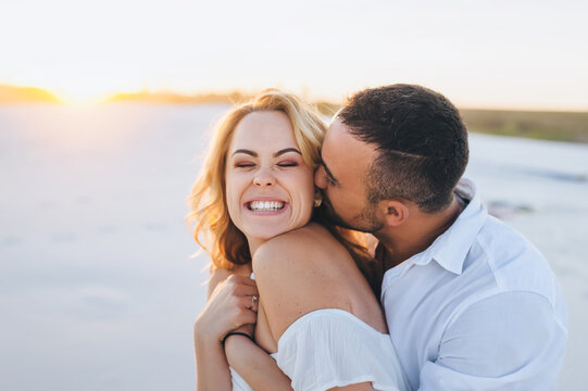 A tanned caucasian bearded guy in a white shirt kisses a young beautiful cheerful smiling blonde against the backdrop of the setting sun. Desert, sandy beach, rest and relaxation. Honeymoon concept.