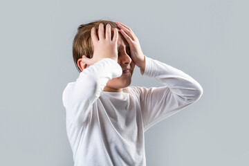 Little boy having a headache. Despair, tragedy. Headache child. Suffering migraine. Headache because stress. Portrait of a sad boy holding his head with his hand, isolated on the gray background