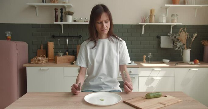 Woman at kitchen looking sad and bored with diet not wanting to eat slice of cucumber. Healthy nutrition and dieting eating
